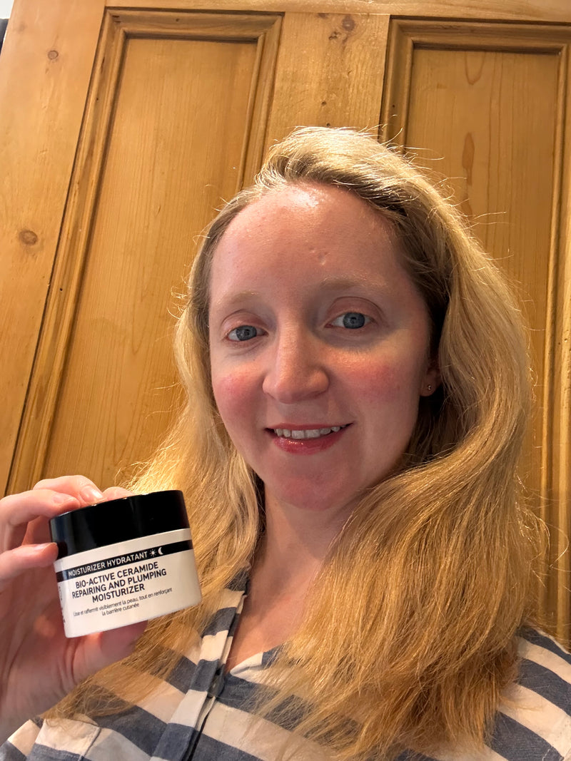 A woman with blonde hair holds a jar of moisturiser in front of a wooden door background.