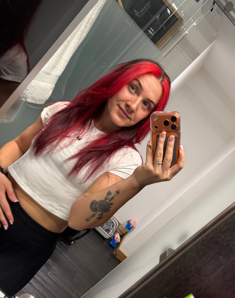 A woman with vibrant red and black hair takes a mirror selfie wearing a white crop top and black pants, holding a brown smartphone.