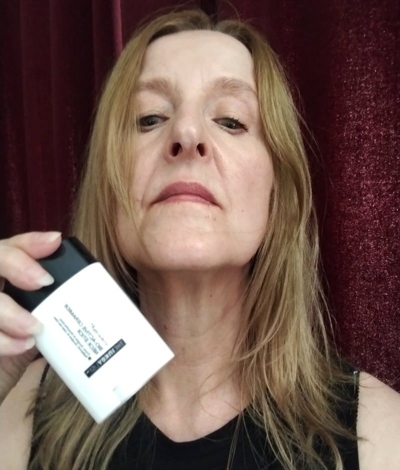 A woman holding a white and black cosmetic bottle in front of a dark red curtain background.