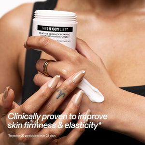 A close-up of a person applying white cream from The Inkey List Bioactive Moisturizer onto their hand. The person is wearing beige manicured nails and gold rings, and the background is a soft-focus view of their bare torso.