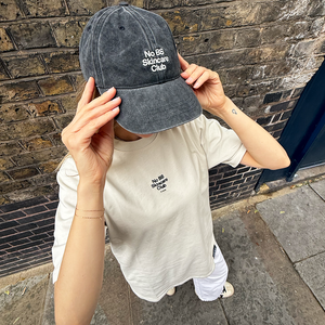 Person wearing a cap and t-shirt with 'No BS Skincare Club' text against a brick wall.