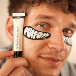 Man holding a tube of 'Puff Off' caffeine eye cream with a black eye patch on his face.