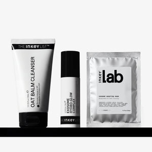 Three skincare products from The Inkey List arranged on a black shelf against a light gray background: a oat Balm Cleanser tube, an Exosome Complex bottle, and an Exosome Boosting Mask sachet.