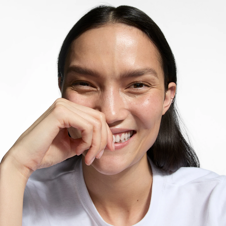 A close-up portrait of a smiling woman with dark hair wearing a white crew-neck t-shirt, set against a plain white background.