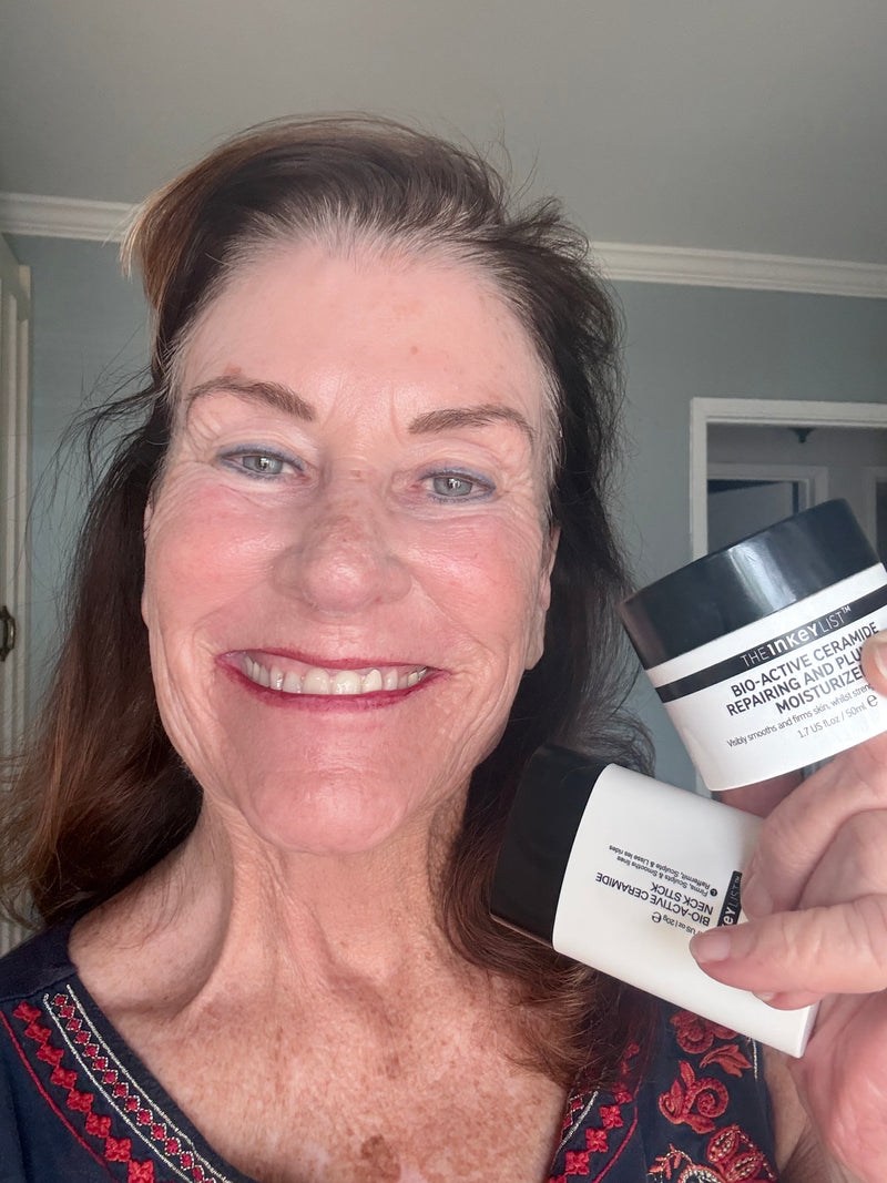 A smiling woman holds two skincare products in front of her face: a white jar labeled Ceramide Restoring Moisturizer' and a white stick labeled 'Bio-Active Moisturizer'. The photo is taken indoors with a light gray wall and doorway in the background.