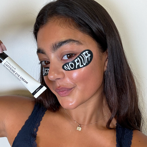 Woman holding a tube of caffeine eye cream with 'NO FLUFF' eye patch under her eye.