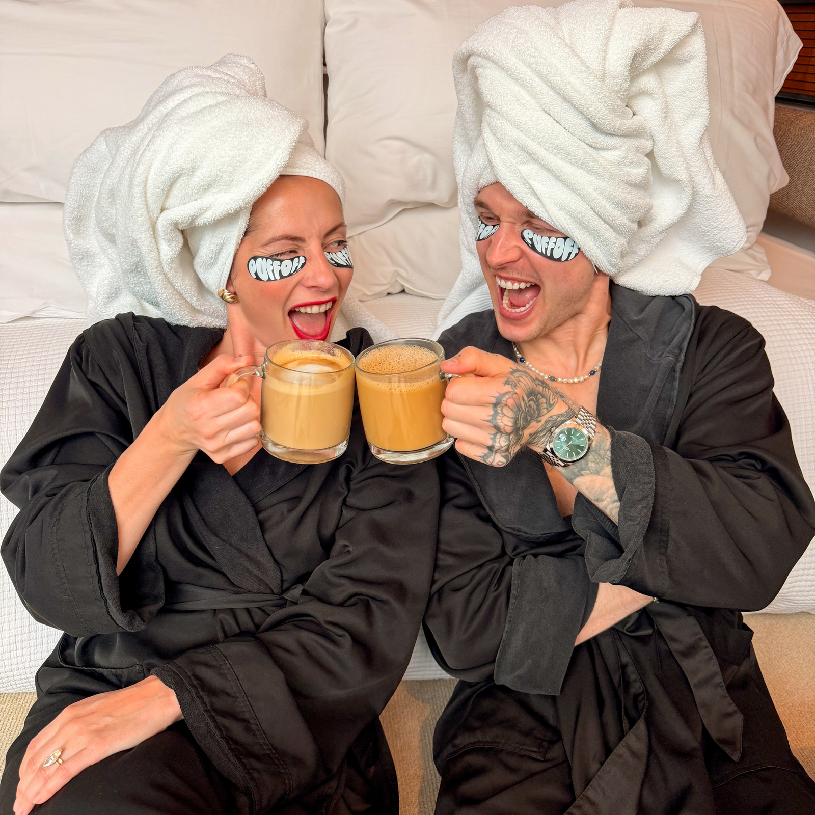 Two people in black robes with towels on their heads, holding coffee mugs and smiling.