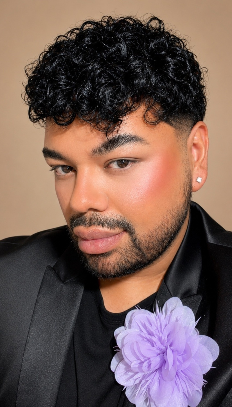 A close-up portrait of a man with curly black hair wearing a black satin jacket, black shirt, and a large light purple flower boutonniere against a warm beige background.