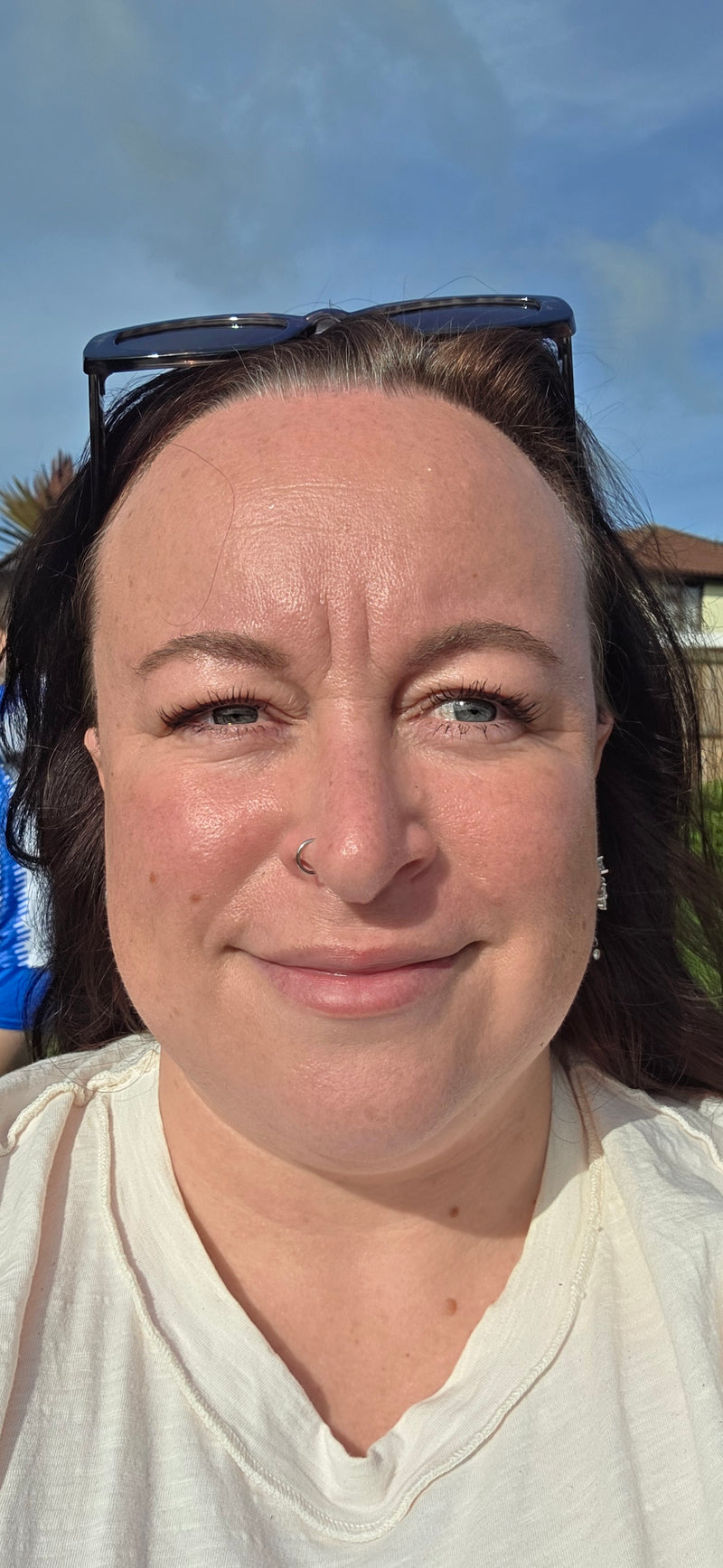 A close-up selfie of a woman outdoors under a blue sky, wearing a white t-shirt and sunglasses on her head.