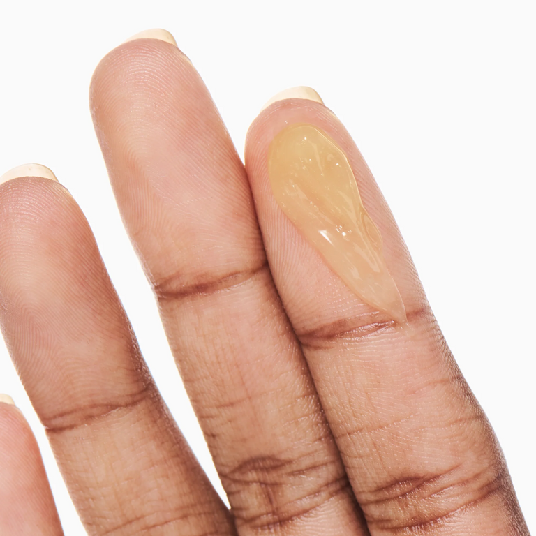 A close-up of a person's fingers showing a glossy, translucent beige gel or cream swatch on the middle finger against a plain white background.