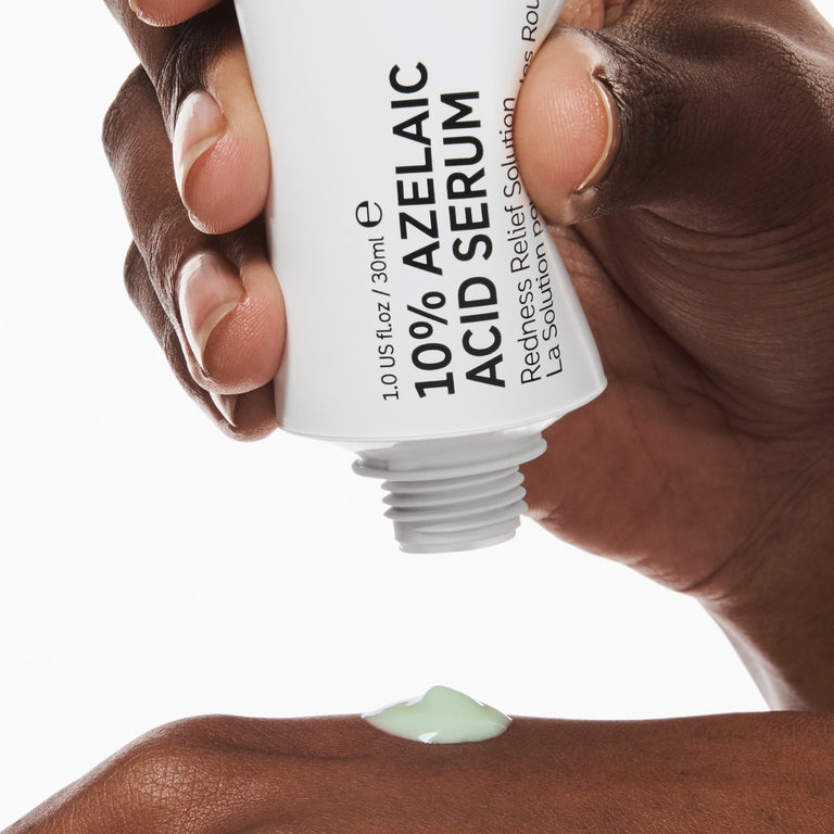 A close-up of a hand squeezing a white tube of 10% Azelaic Acid Serum, with a small dollop of pale green serum dripping onto another hand against a white background.