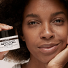 A close-up lifestyle shot of a woman with warm skin holding a black jar Moisture Cream against a dark background.
