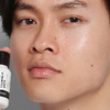 Close-up of a man's face with a glossy, hydrated finish, holding a black cosmetic bottle with white text near his cheek against a neutral gray background.