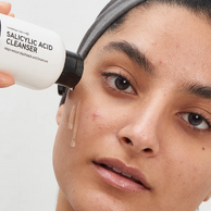 A close-up of a woman applying or demonstrating a skincare cleanser, with a hand holding a white bottle labeled 'SALICYLIC ACID CLEANSER' against a clean light background.