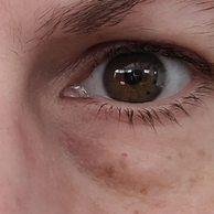 Extreme close-up of a human eye and surrounding skin, showing a brown iris, eyelashes, eyebrow, and under-eye area.