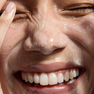 Extreme close-up of a person's face with white powder or sugar sprinkled across the skin, showing a wrinkled, wrinkled nose, closed eyes, and teeth.