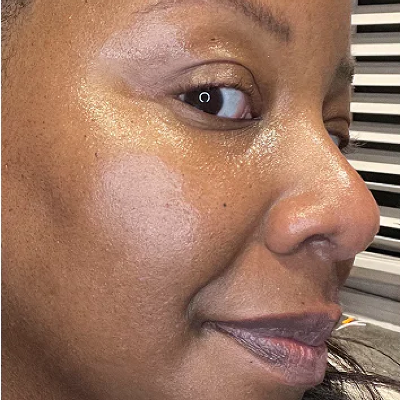 Close-up of a woman's face showing a glossy, dewy complexion with a shimmering finish, likely demonstrating a skincare or makeup product.