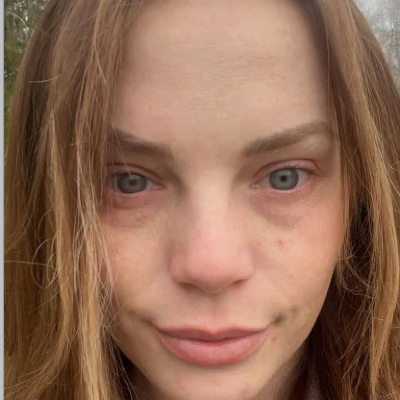A close-up portrait of a young woman with light brown hair, freckles, and blue eyes looking directly at the camera.