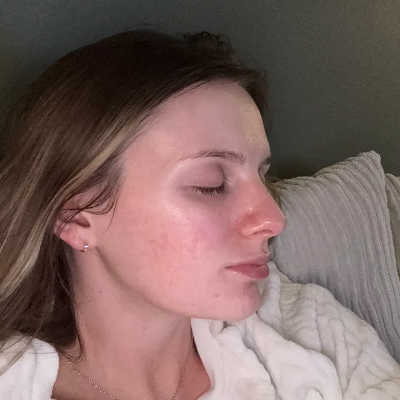 A close-up profile shot of a woman with closed eyes, wearing a small stud earring and a delicate necklace, resting against a textured pillow and white blanket.