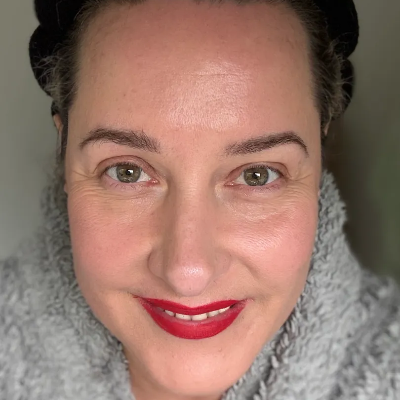 Close-up portrait of a woman with natural skin texture, wearing a gray fuzzy knit top and red lipstick, with her hair pulled back.