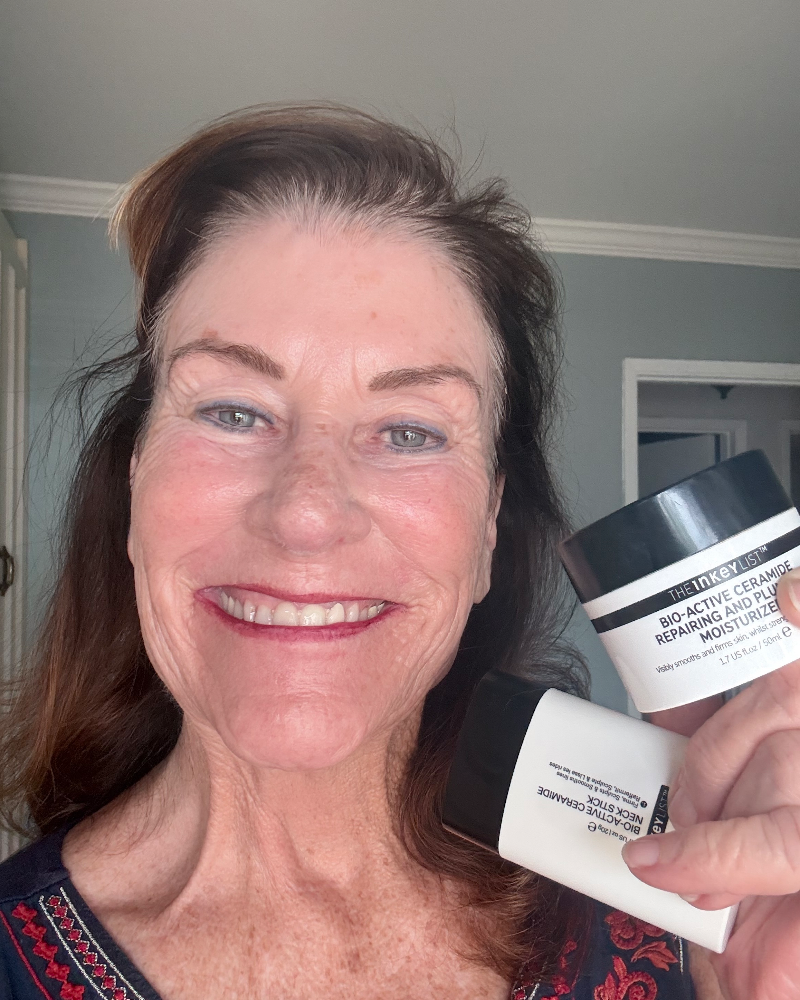 A smiling woman holds two skincare products in front of her face: a white jar labeled The Trudon Bio-Acetician Cleansing & Moisturizing Mask and a white tube labeled The Trudon Skin Support Serum.