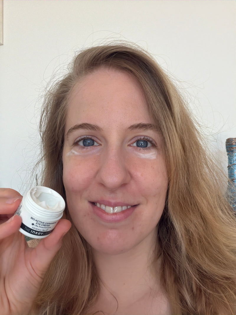 A woman holding a small white jar of skincare cream next to her face, which has matching white cream applied under her eyes. She is smiling at the camera against a plain off-white wall.