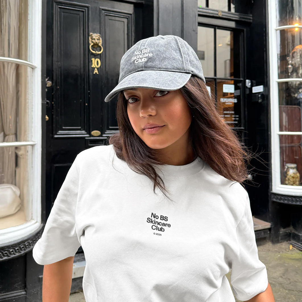 Person wearing a white t-shirt and black cap with text, standing in front of a building entrance.